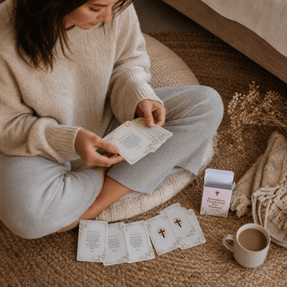 A woman sits cross-legged on a woven rug, reading cards from a deck. The cards are shown both in her hands and arranged on the floor in front of her.
